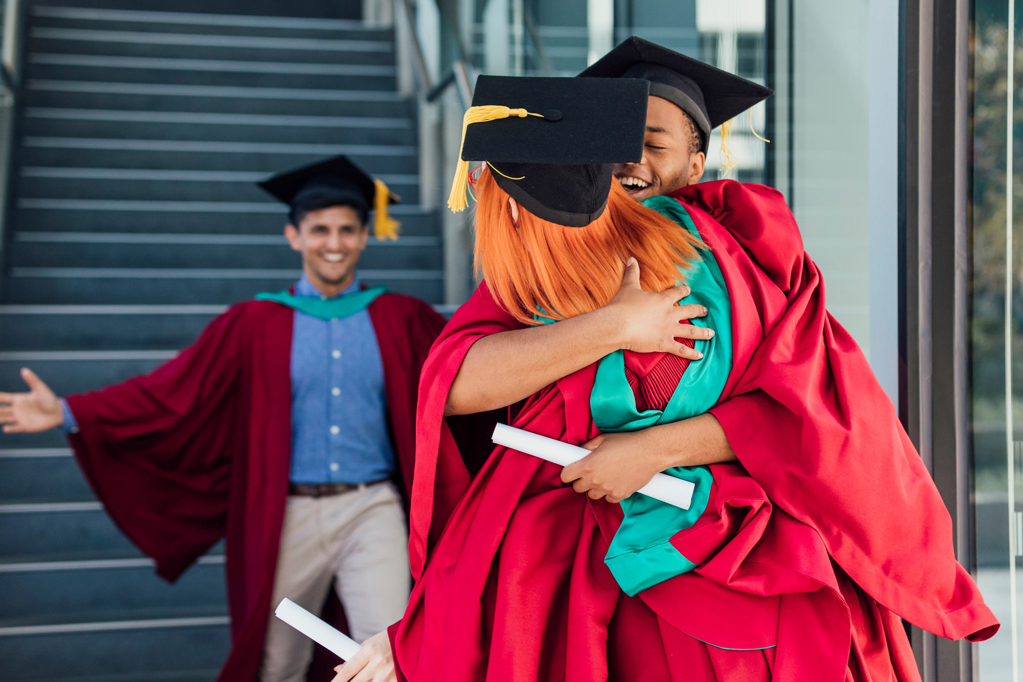 Young people in graduation gowns and hats hugging outside building